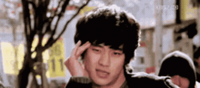 A Young Man Is Holding His Head In Pain While Standing In Front Of A Tree . GIF