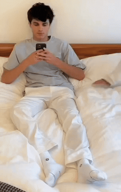 A Young Man Is Laying On A Bed Looking At His Cell Phone GIF