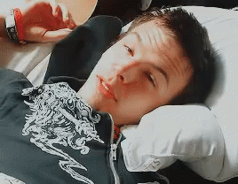 A Young Man Is Laying On A Bed With His Head On A Pillow And Wearing A Black Shirt With A Dragon On It GIF