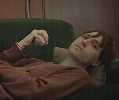 A Young Man Is Laying On A Green Couch With His Hand On His Face GIF