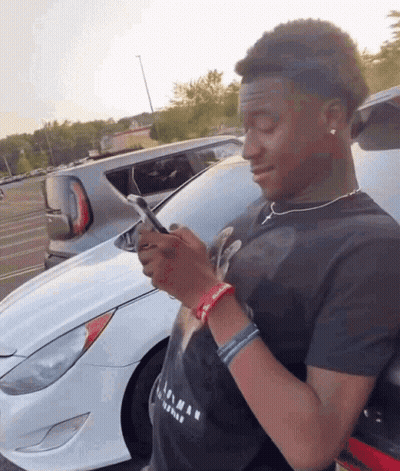 A Young Man Is Looking At His Phone In Front Of A Car GIF