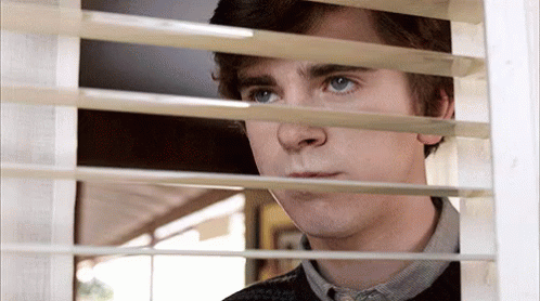 A Young Man Is Looking Out Of A Window With Blinds . GIF
