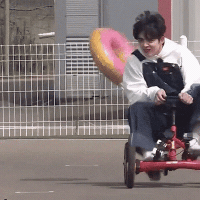 A Young Man Is Riding A Tricycle With A Pink Donut In The Back . GIF