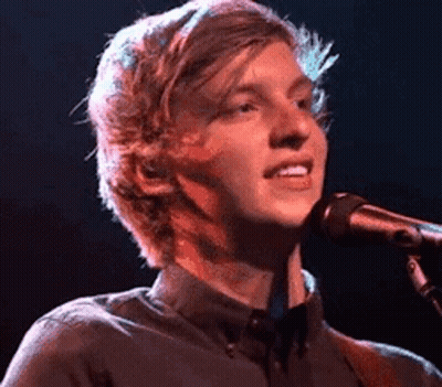 A Young Man Is Singing Into A Microphone While Playing A Guitar On Stage . GIF