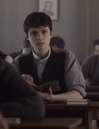 A Young Man Is Sitting At A Desk In A Classroom Holding A Book GIF