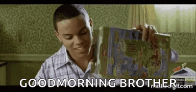 A Young Man Is Sitting At A Table Holding A Box Of Cereal And Smiling . GIF