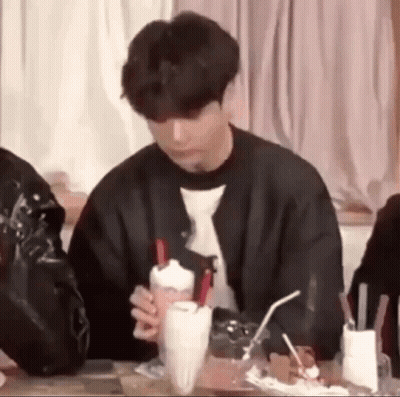 A Young Man Is Sitting At A Table Holding A Milkshake With Straws . GIF