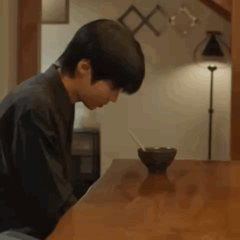 A Young Man Is Sitting At A Table With A Bowl Of Food And A Spoon In It . GIF