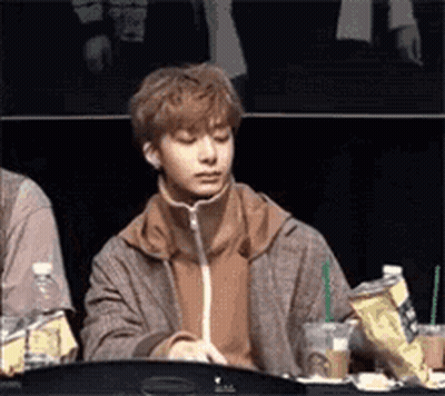 A Young Man Is Sitting At A Table With A Drink In His Hand And A Bag Of Chips . GIF