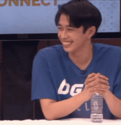 A Young Man Is Sitting At A Table With His Hands Folded And Smiling . GIF