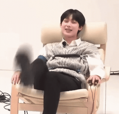 A Young Man Is Sitting In A Chair With His Legs Crossed And A Sweater On . GIF
