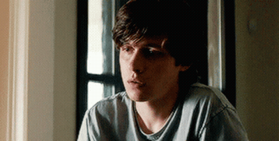 A Young Man Is Sitting In Front Of A Window Wearing A Grey Shirt . GIF