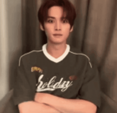 A Young Man Is Sitting In Front Of A Window With His Arms Crossed And Wearing A Holiday Shirt . GIF