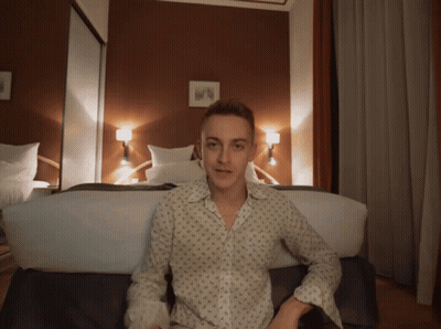 A Young Man Is Sitting On A Bed In A Bedroom GIF