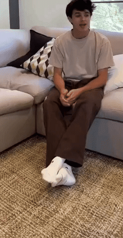 A Young Man Is Sitting On A Couch Wearing White Socks And A White Shirt . GIF