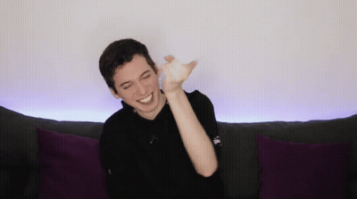 A Young Man Is Sitting On A Couch With Purple Pillows And Smiling GIF