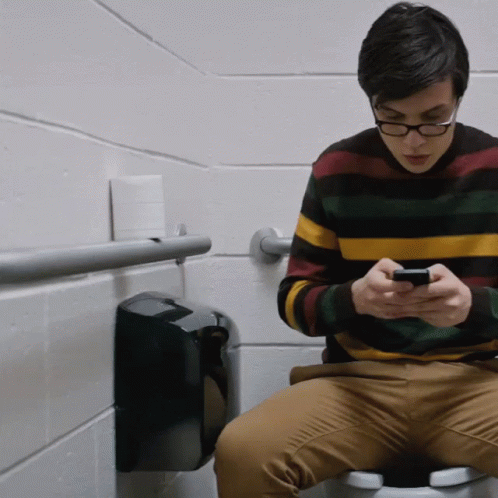 A Young Man Is Sitting On A Toilet Looking At His Phone GIF