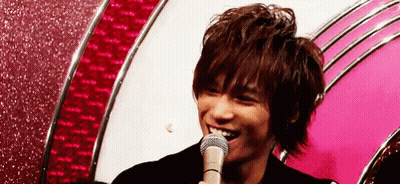 A Young Man Is Smiling While Holding A Microphone In Front Of A Pink Wall . GIF