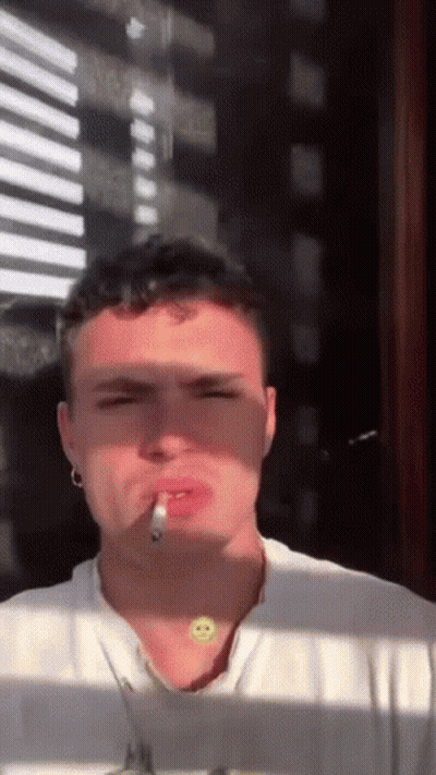 A Young Man Is Smoking A Cigarette In Front Of A Window While Wearing A Smiley Face Necklace . GIF