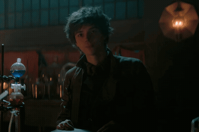 A Young Man Is Standing In A Dark Room In Front Of A Beaker With Blue Liquid In It . GIF