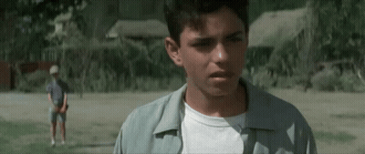 A Young Man Is Standing In A Field Looking At The Camera While Another Young Man Stands In The Background . GIF