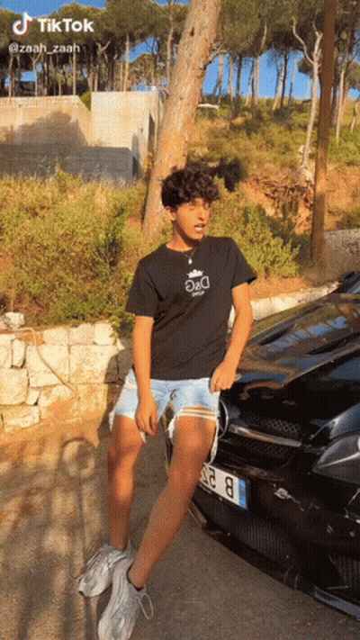 A Young Man Is Standing In Front Of A Black Car . GIF