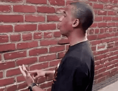 A Young Man Is Standing In Front Of A Red Brick Wall And Pointing At It . GIF