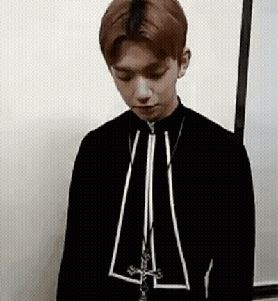 A Young Man Is Standing In Front Of A White Wall Wearing A Black Jacket And A Necklace . GIF