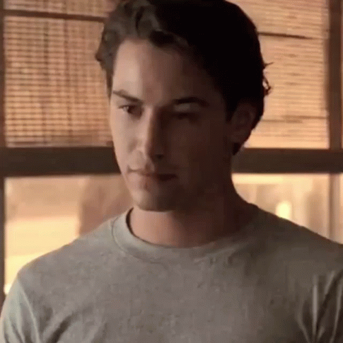 A Young Man Is Standing In Front Of A Window Wearing A Grey Shirt . GIF