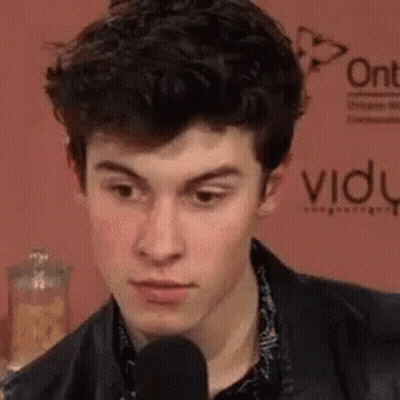 A Young Man Is Talking Into A Microphone While Wearing A Black Jacket . GIF