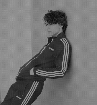 A Young Man Leaning Against A Wall Wearing An Adidas Tracksuit GIF