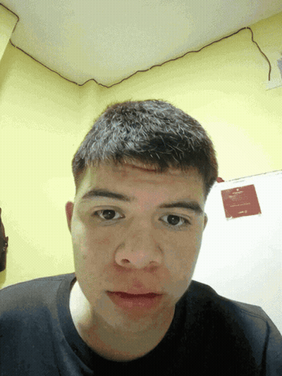 A Young Man Looks At The Camera In Front Of A Yellow Wall With A Sign That Says " No Smoking " On It GIF