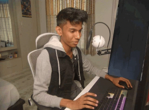 A Young Man Sits At A Desk With A Computer And A Microphone GIF