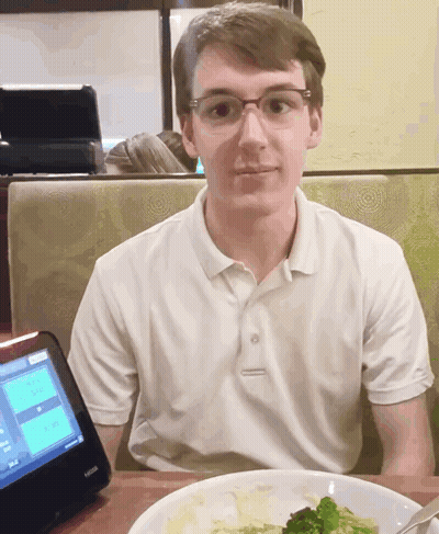 A Young Man Sits At A Table In Front Of A Kiosk That Shows A Menu GIF