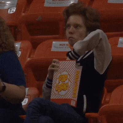 A Young Man Sitting In A Stadium Holding A Box Of Pop Corn GIF