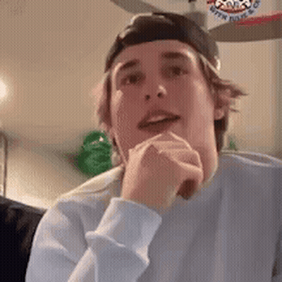 A Young Man Wearing A Baseball Cap And A White Sweater Is Sitting In A Living Room With His Hands On His Chin . GIF
