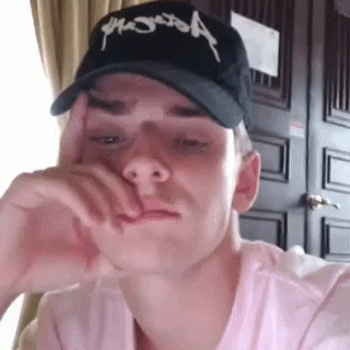 A Young Man Wearing A Baseball Cap Is Sitting In Front Of A Door . GIF