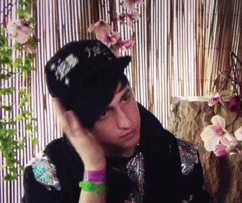 A Young Man Wearing A Black Hat With Chinese Characters On It Is Sitting In Front Of Flowers GIF