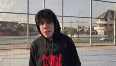 A Young Man Wearing A Black Hoodie And A Red Shirt Is Standing In Front Of A Fence . GIF
