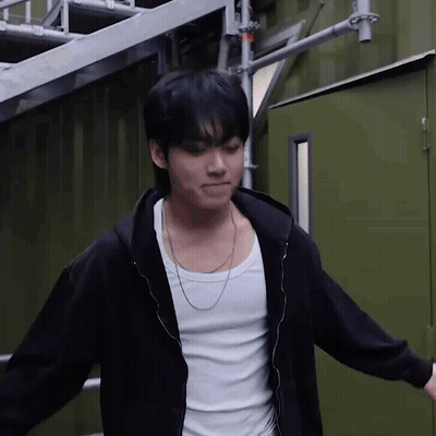 A Young Man Wearing A Black Hoodie And A White Tank Top Is Standing In Front Of A Green Building . GIF