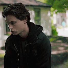 A Young Man Wearing A Black Jacket And A Black Hoodie Is Standing In Front Of A House . GIF