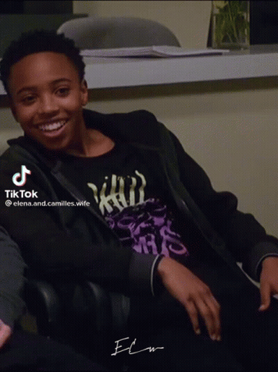 A Young Man Wearing A Black Jacket And A Purple Shirt Is Smiling And Sitting In A Chair GIF