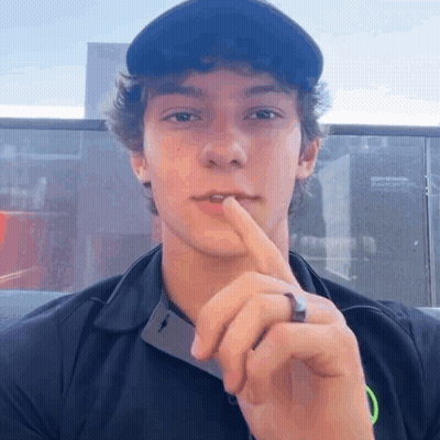 A Young Man Wearing A Black Shirt And A Black Hat Holds His Finger To His Lips GIF