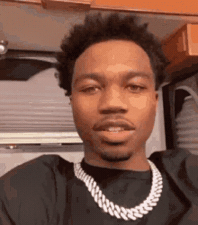 A Young Man Wearing A Black Shirt And A Chain Around His Neck Is Looking At The Camera . GIF