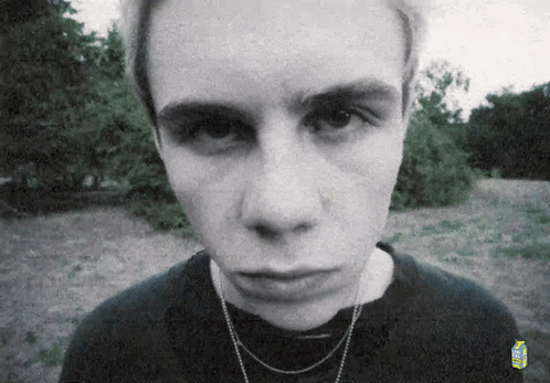 A Young Man Wearing A Black Shirt And A Necklace Looks At The Camera . GIF