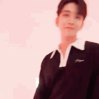 A Young Man Wearing A Black Sweater And A White Collar Is Standing In Front Of A White Wall . GIF