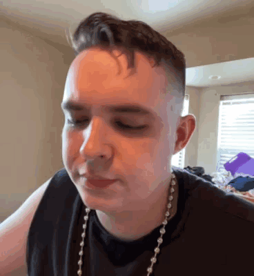 A Young Man Wearing A Black Tank Top And A Pearl Necklace Is Making A Face . GIF