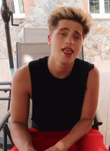 A Young Man Wearing A Black Tank Top And Red Shorts Is Sitting In A Chair GIF