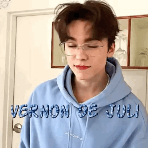 A Young Man Wearing A Blue Hoodie And Glasses Is Standing In Front Of A Door . GIF