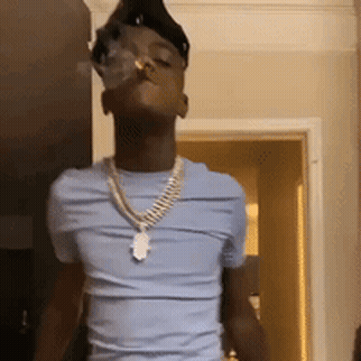 A Young Man Wearing A Blue Shirt And A Gold Chain Is Smoking A Cigarette In A Room . GIF